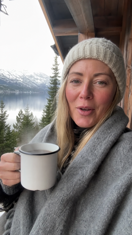 AI-generated creator image: a woman wrapped in a wool blanket on a cabin porch holding a mug toward the phone, fjord and snow behind