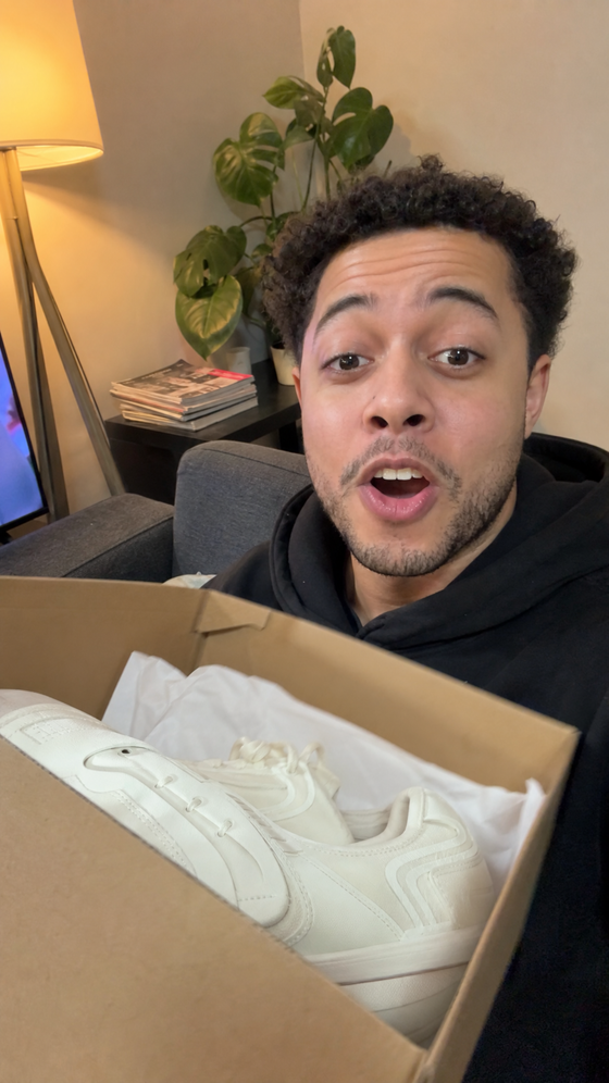AI-generated creator image: a man on his sofa holding a sneaker box open toward the phone camera, excited, evening lamp light