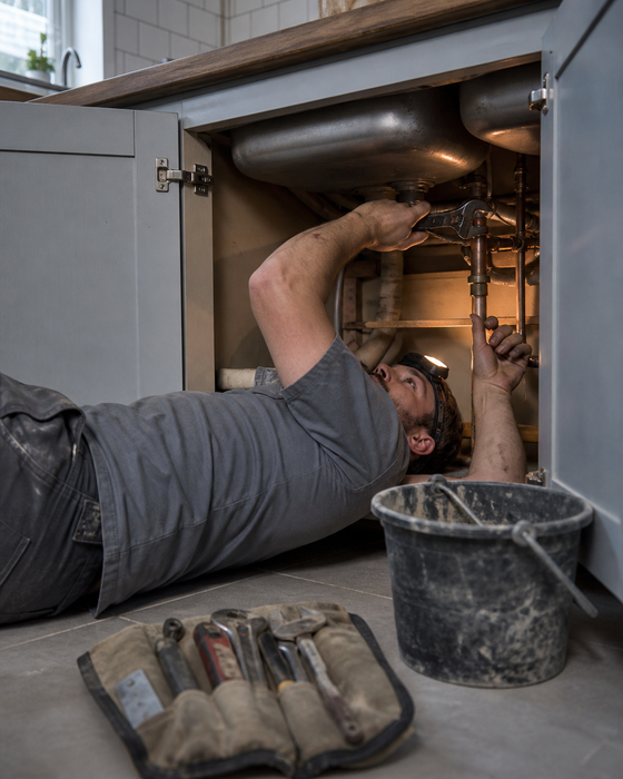 AI-generated ad image: a plumber tightening a copper fitting under a kitchen sink, torch light, tools on the floor