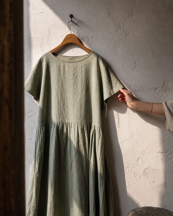 AI-generated ad image: a sage linen dress on a wooden hanger against a plaster wall, sunlight patch, a woman's hand touching the fabric